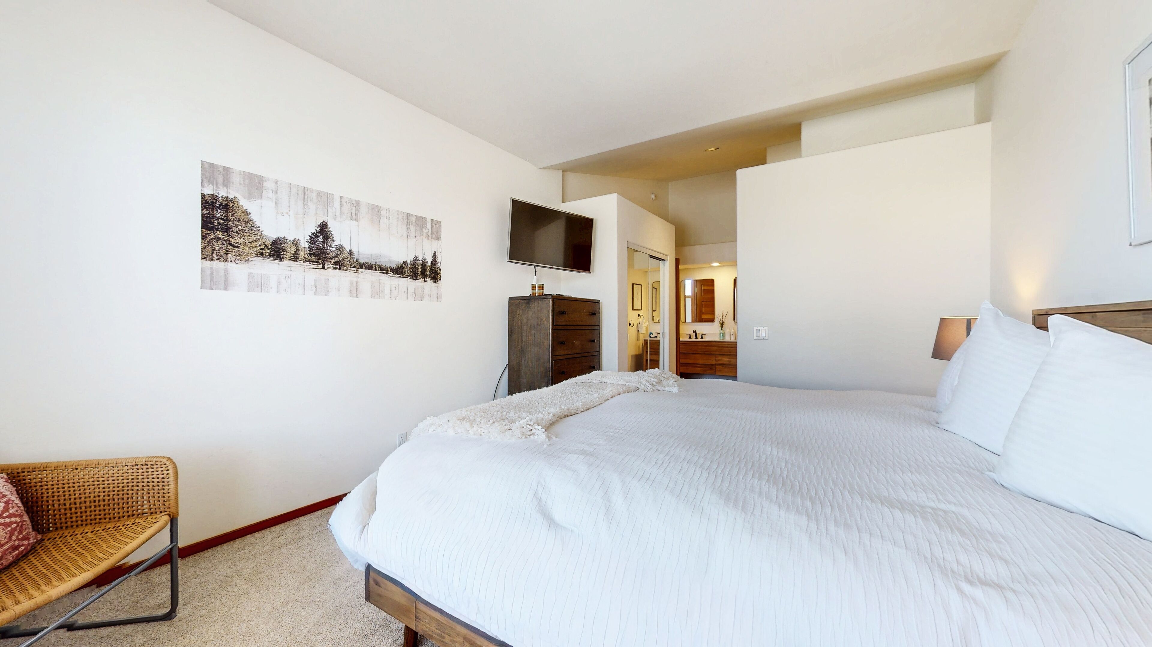 Une chambre à coucher avec un grand lit, une commode en bois, un téléviseur fixé au mur et un tableau représentant un paysage enneigé.