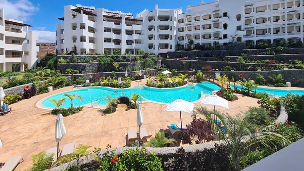 A hotel with a swimming pool surrounded by lounge chairs and umbrellas, set against a backdrop of white buildings and greenery. - BEAUTIFUL APARTMENT IN RESIDENCE JARDINES DE LOS MENCEYES HEATED SWIMMING POOL (Palm-Mar)