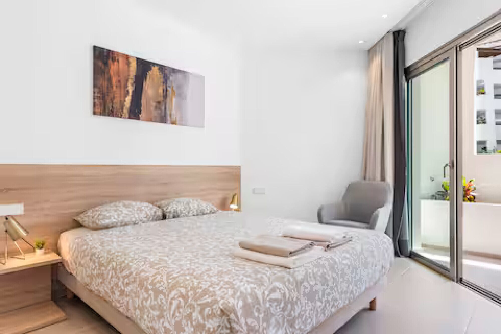 Une chambre moderne avec un grand lit, une tête de lit en bois, une table de chevet avec une lampe, une chaise et une grande fenêtre donnant sur des immeubles.