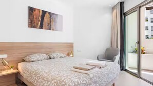 A modern bedroom with a large bed, a wooden headboard, a bedside table with a lamp, a chair, and a large window with a view of buildings. - BEAUTIFUL APARTMENT IN RESIDENCE JARDINES DE LOS MENCEYES HEATED SWIMMING POOL (Palm-Mar)