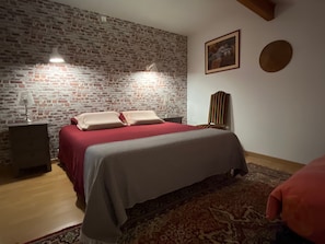 A bedroom with a brick wall, a bed with red bedding, two bedside lamps, a chair, a rug, and a painting on the wall. - Léontine Guest Room – Canal des Deux Mers by Bike (Lamagistère)