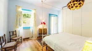 Une chambre à coucher avec un lit, deux chaises en bois, une petite table avec une lampe et une fenêtre avec des rideaux.