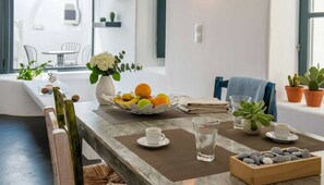 A dining area with a wooden table, a vase of flowers, a fruit bowl, and a tray of bread. - Dionysos Wine Authentic Stay 2 Bd Santorini Home (Santorini)
