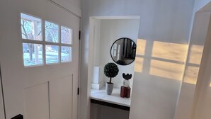 A hallway with a white door, a window, a small shelf with a potted plant, a mirror, and a wall-mounted speaker.