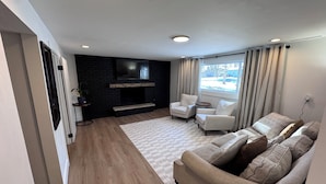 A modern living room with a fireplace, two white armchairs, a beige sofa, and a large window with curtains.