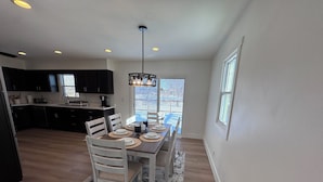 A dining area with a table set for four, surrounded by white chairs, a black ceiling light, and a view of the outdoors through a window.