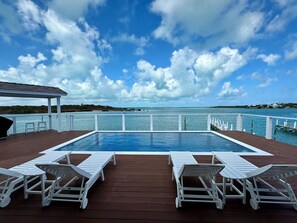 A pool area with white lounge chairs, a wooden deck, and a view of the sea and distant land under a partly cloudy sky. - Three Little Birds: Waterfront Home-Private Pool & Dock Perfect For Boaters! (Hope Town)