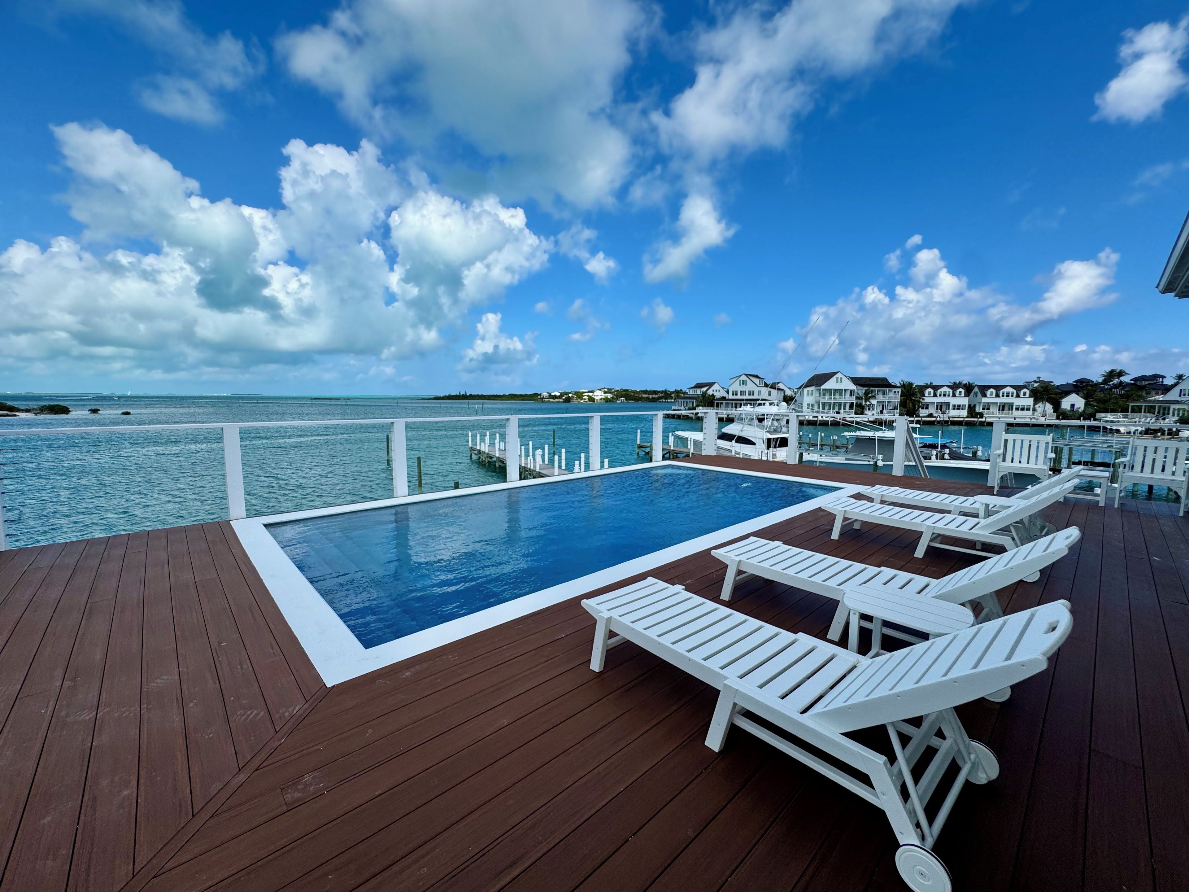 A wooden deck with a pool and lounge chairs overlooking a marina and ocean.