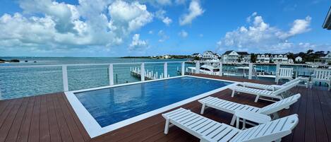 A wooden deck with a pool and lounge chairs overlooking a marina and ocean.