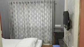 Un dormitorio con cama, aire acondicionado, televisor montado en la pared, cortina y un cubo de basura azul pequeño.