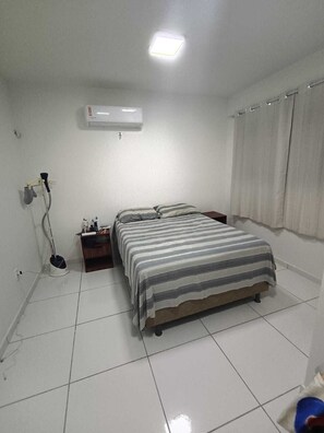 Een slaapkamer met een bed, nachtkastje, een wandairconditioner, raam met gordijnen en een vloerlamp.