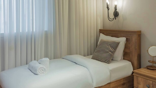 Una cama bien hecha con un edredón blanco, una almohada gris y una toalla blanca enrollada. Hay una mesita de noche de madera con un espejo redondo y un cajón pequeño. La habitación tiene un cabecero de madera y una lámpara de pared.