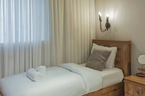 Een net opgemaakt bed met een witte dekbedovertrek, een grijs kussen en een opgerolde witte handdoek. Er staat een houten nachtkastje met een rond spiegelbeeld en een klein lade. De kamer heeft een houten hoofdbord en een wandlamp.
