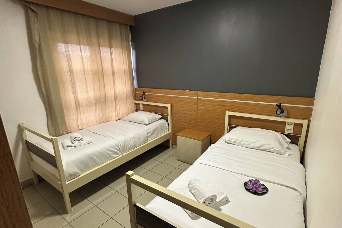 Une chambre d’hôtel avec deux lits simples, une tête de lit en bois, une petite table de chevet et un luminaire au plafond.