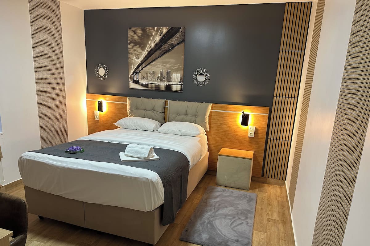 Une chambre d’hôtel moderne dotée d’un grand lit, de tables de chevet, d’une tête de lit avec éclairage intégré, d’une œuvre d’art représentant un paysage urbain au-dessus du lit et d’un panneau mural à motifs.