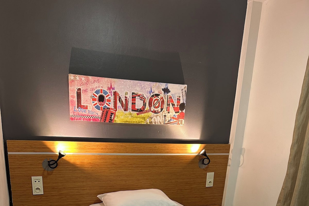 Une chambre d’hôtel avec un lit, une tête de lit en bois, une enseigne fixée au mur sur laquelle on peut lire « LONDRES », et une seule fleur sur le lit.