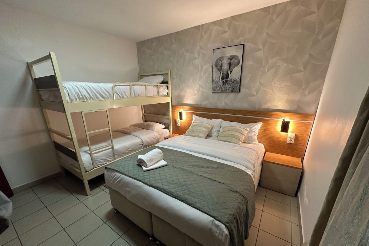 Une chambre d’hôtel avec des lits superposés, un grand lit recouvert d’une courtepointe matelassée, un tableau représentant un éléphant accroché au mur et un motif texturé sur le mur.