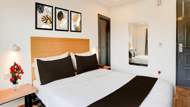 Ein Hotelzimmer mit einem Bett, einem Nachttisch mit Blumen in einer Vase, einem Wandspiegel und drei gerahmten Kunstwerken über dem Bett.