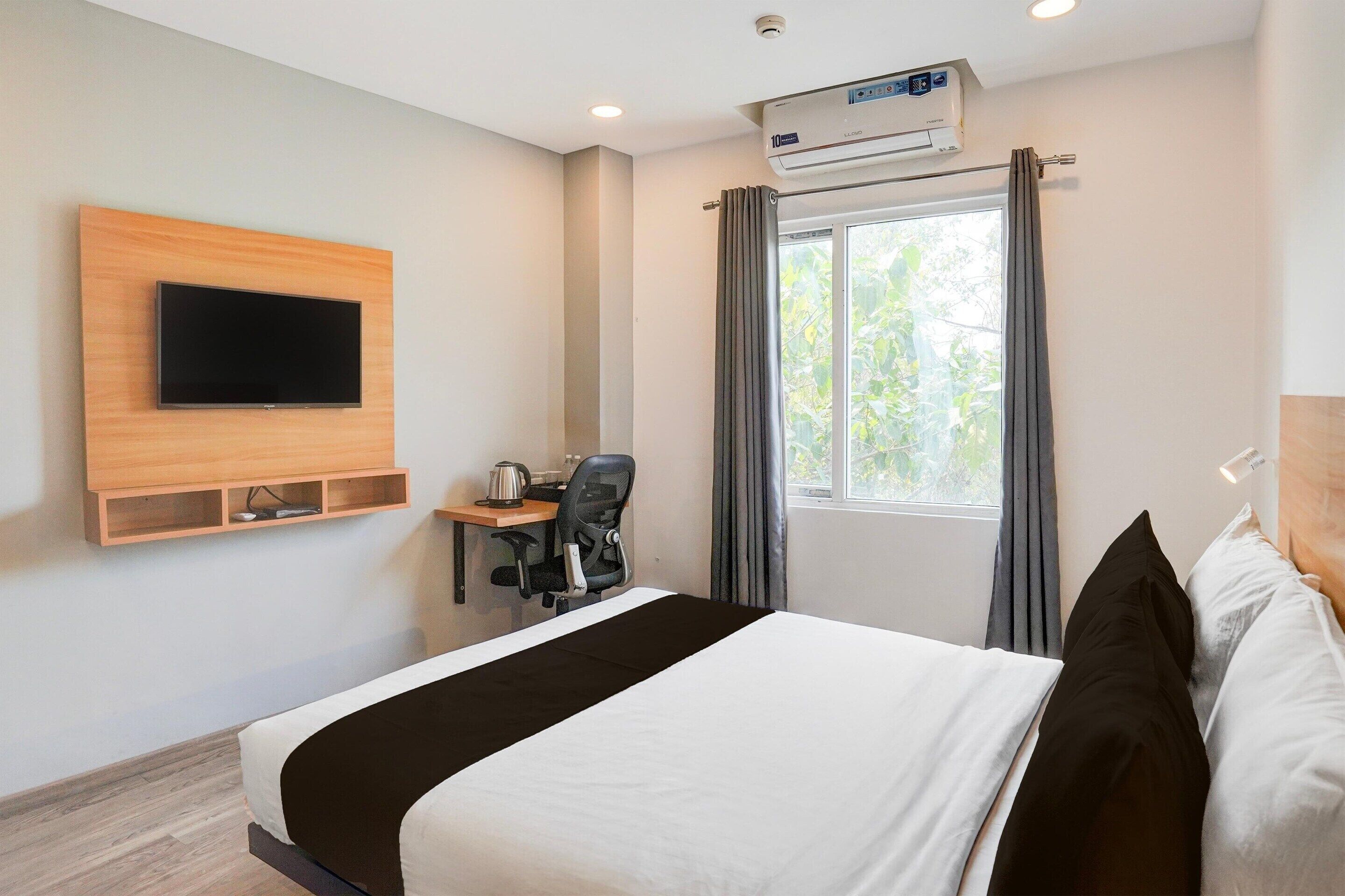 A hotel room with a bed, a desk with a chair, a flat-screen TV mounted on the wall, and an air conditioning unit above the window.