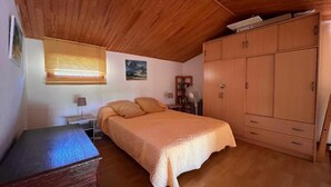 Une chambre avec un plafond en bois, un lit recouvert d’une couverture orange, une armoire en bois et un bureau avec une lampe.