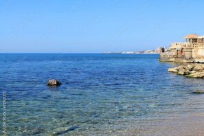 A coastal building with clear blue waters in the foreground and a rocky shoreline.