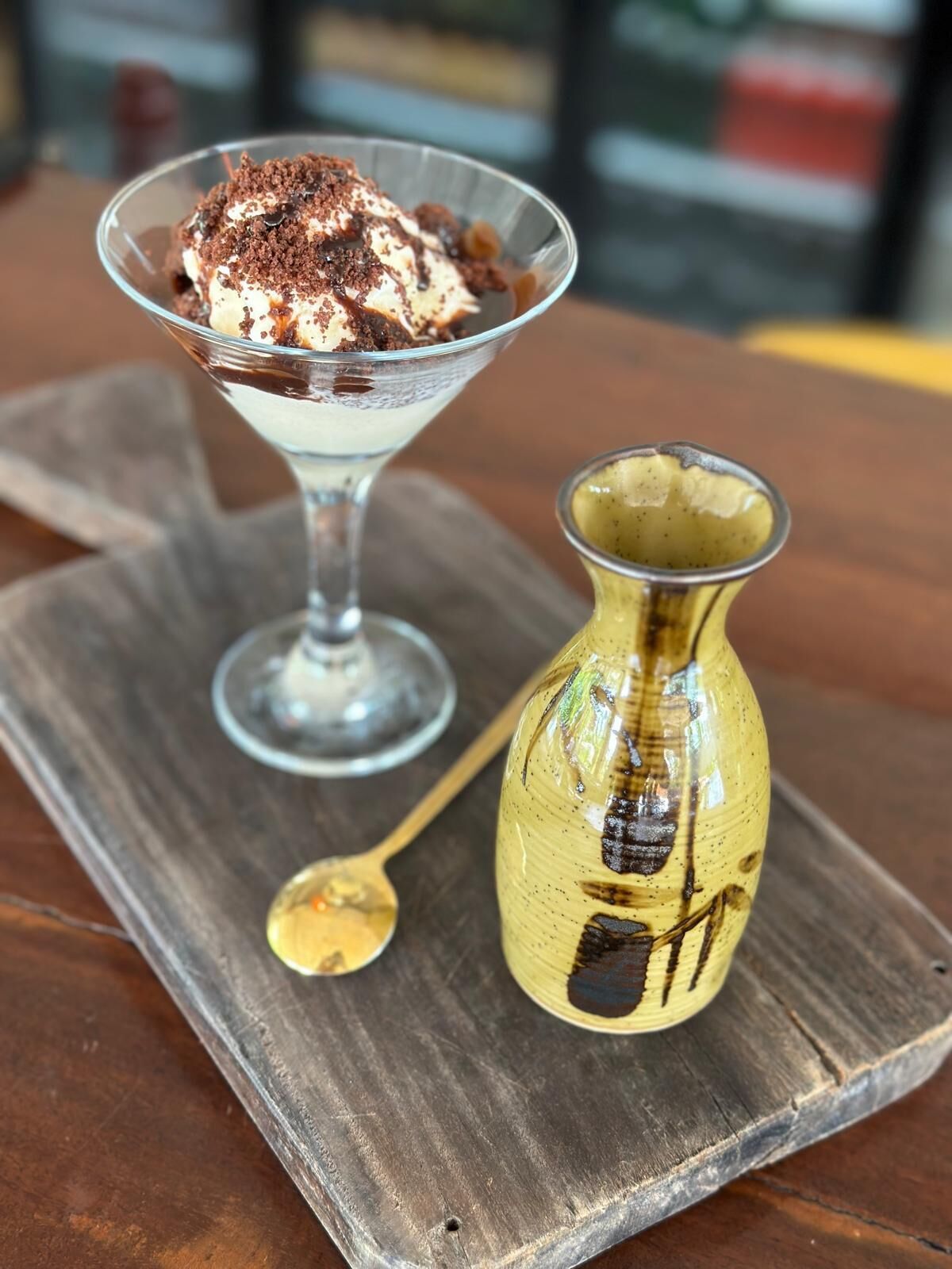 A dessert in a glass bowl on a wooden board, accompanied by a spoon and a decorative vase.