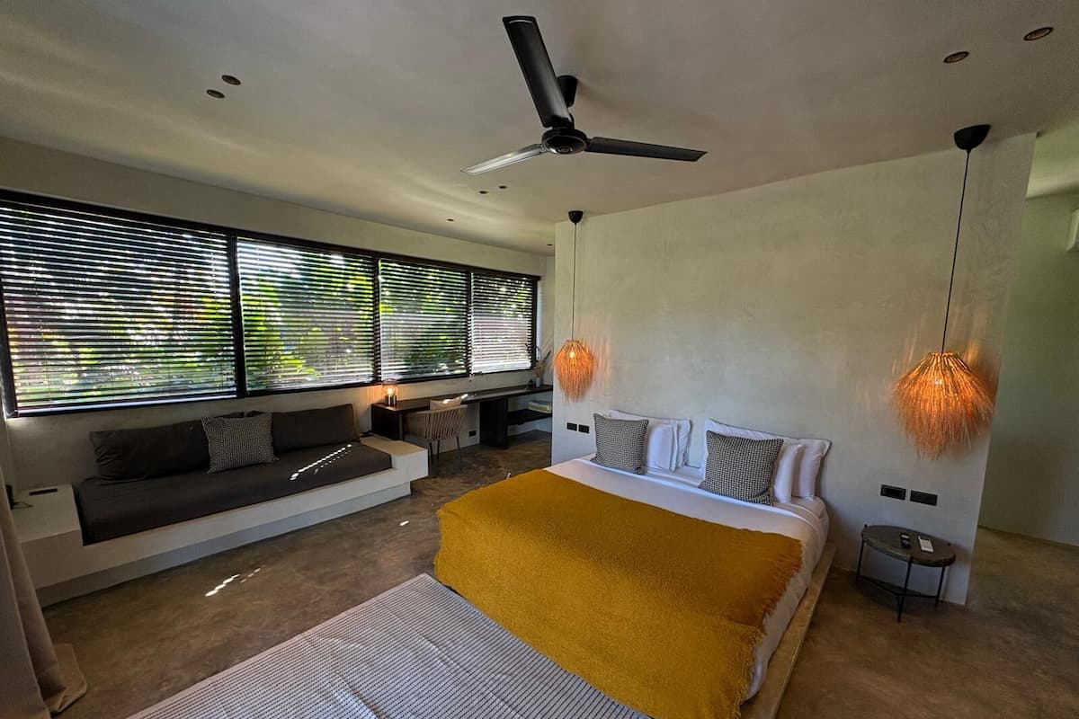 Un dormitorio moderno con cama, sofá y escritorio. Hay ventanas amplias con persianas, un ventilador de techo y luces colgantes.