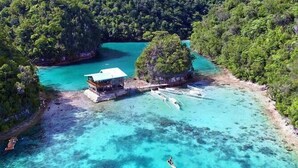 An aerial view of a tropical island with clear turquoise waters, a wooden dock, and a small building. - Surfing Temple Boutique Hotel (General Luna)