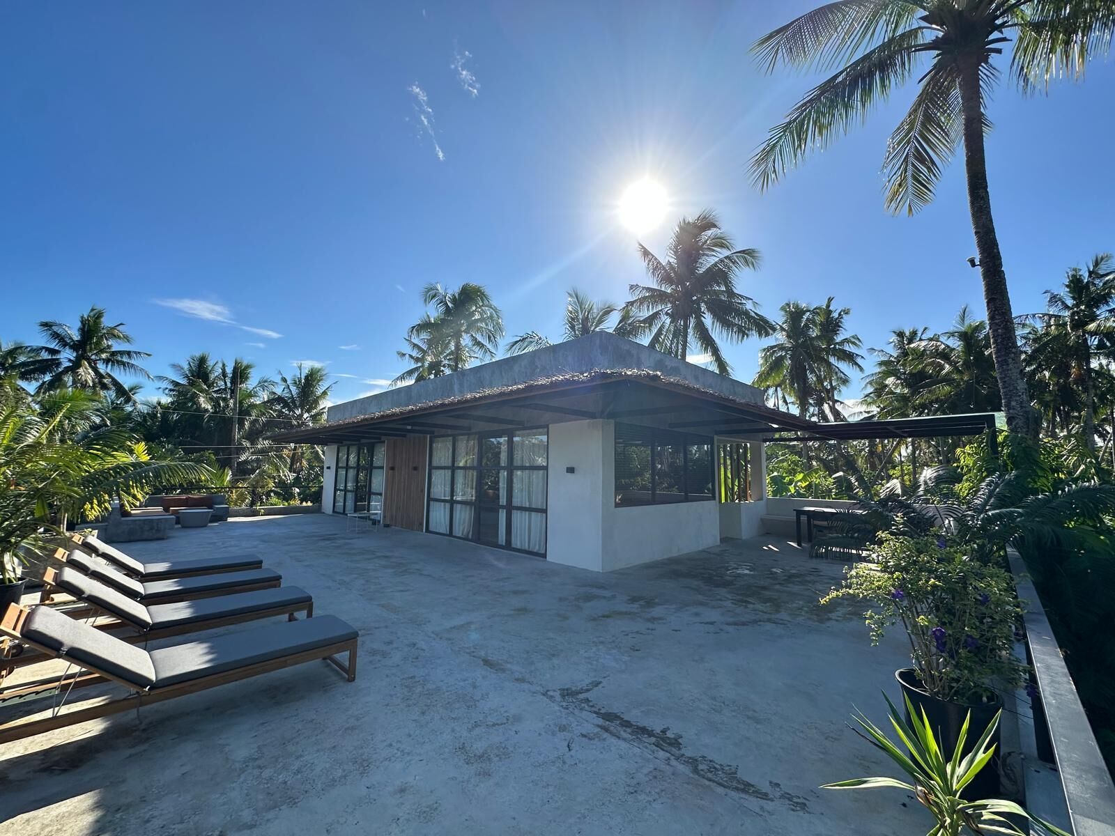 A modern beach house with a covered patio, lounge chairs, and a view of palm trees.
