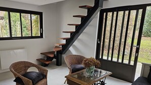 Un interior moderno con una escalera negra, sillas de mimbre, una mesa de centro de vidrio y vistas al exterior a través de grandes ventanales.