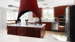 A modern kitchen with a central island, wooden cabinets, stainless steel appliances, and a large window providing natural light. - The Mid-Century Modern House - Long Term Available (Seguin)