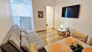 A living room with a sofa, a coffee table with orange placemats, a flat-screen TV mounted on the wall, and a small table with a lamp and remote control. - COOND0124 - Edifício Solarius (Salvador)