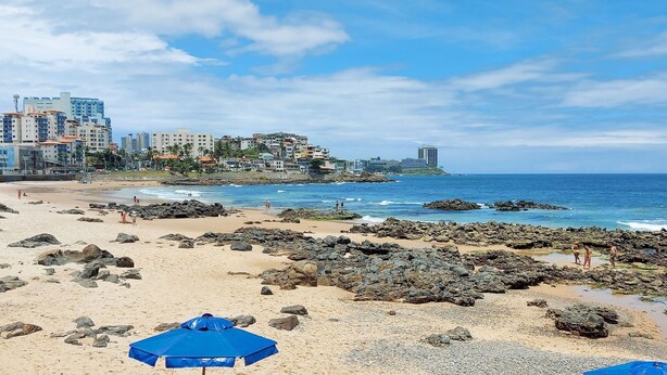 A beach with rocky areas, a few people, and buildings in the background. - COOND0124 - Edifício Solarius (Salvador)