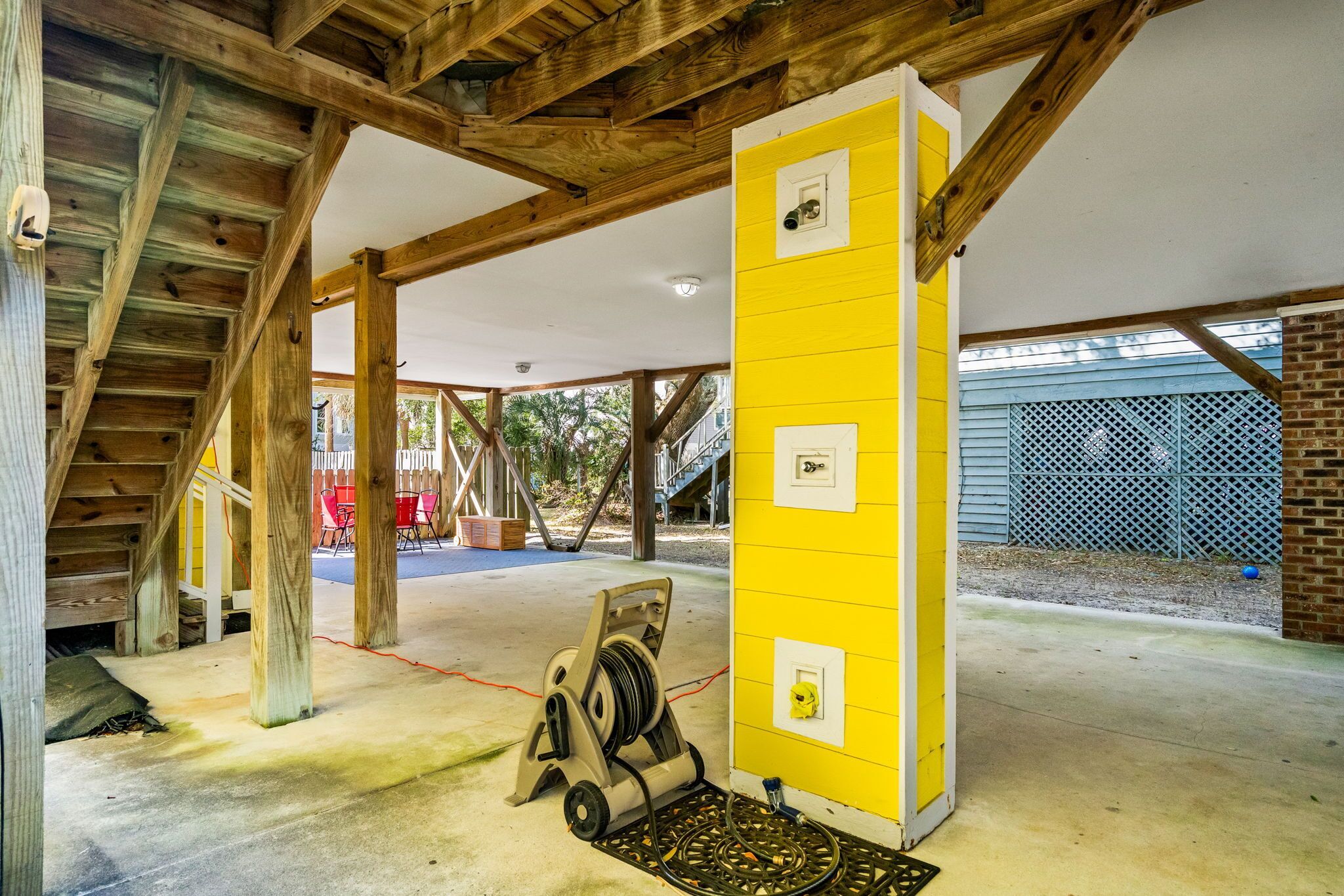 A covered outdoor area with wooden beams, a yellow wall with electrical outlets, and a concrete floor.