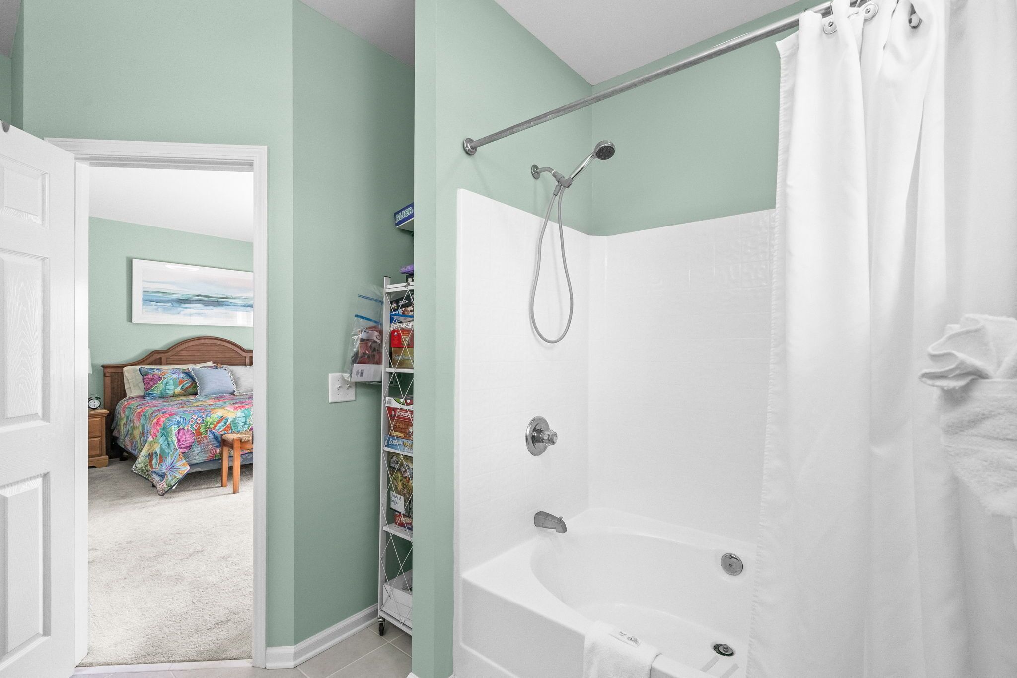 A bathroom with a bathtub, shower, and a door leading to a bedroom with a bed and a bookshelf.