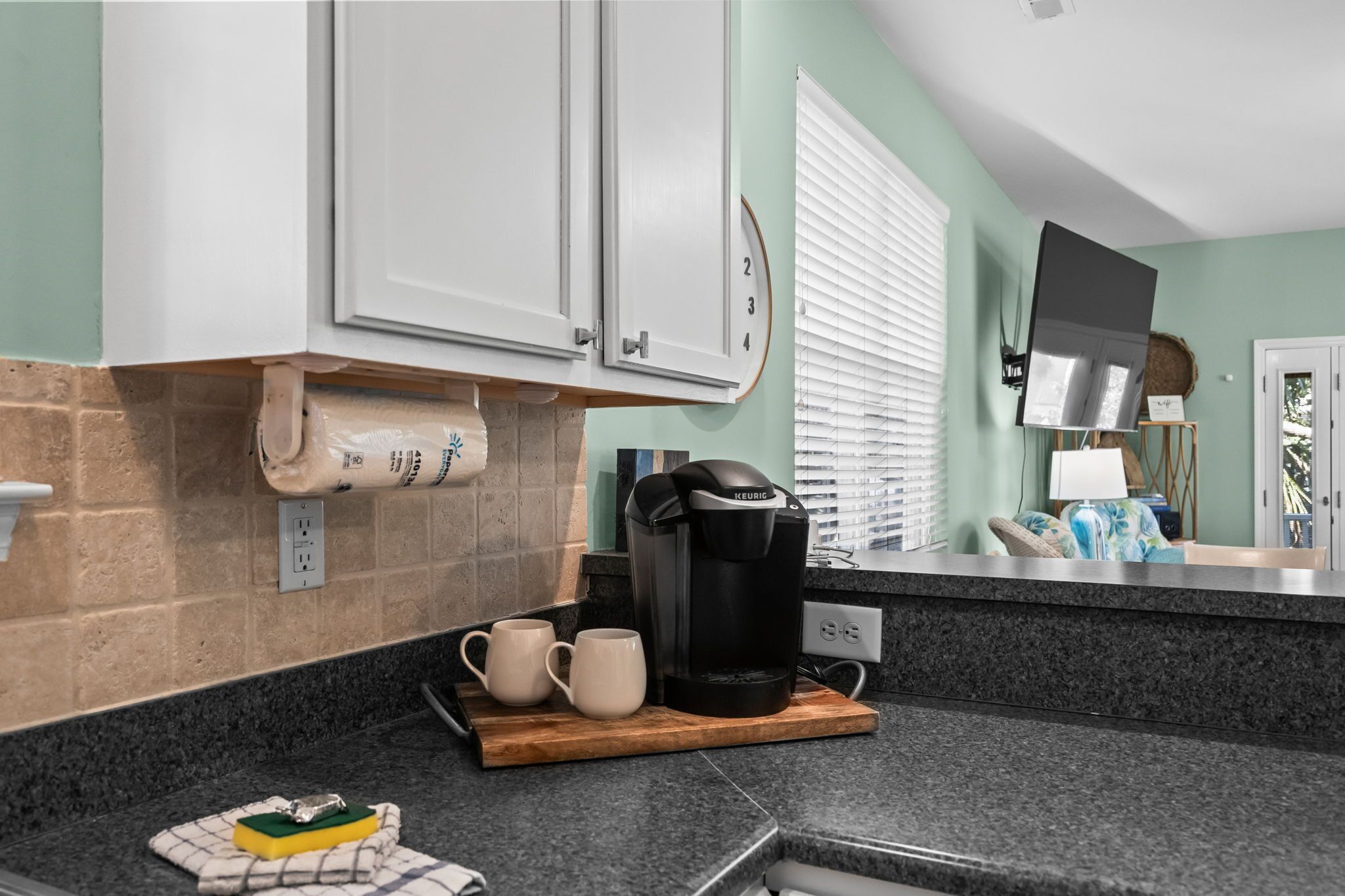 A kitchen with a coffee maker, two mugs, and a sponge on a wooden board.