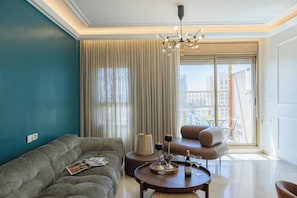 A modern living room with a sofa, armchair, and coffee table, a chandelier, and a view of the city through large windows. - 4 BR Apartment in Sarei Israel Blvd (w/ safe room) (Jerusalem)