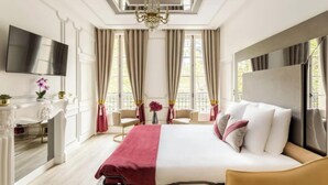 Ein Hotelzimmer mit einem großen Bett, einem an der Wand montierten Fernseher, einem Schreibtisch mit Stuhl und einer Kandelaberleuchte.