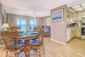 A spacious living area with a dining table, chairs, and a sofa. A kitchen with wooden cabinets and appliances is visible. - Two Bedroom Two Bath Direct Gulf Front Penthouse (Redington Shores)