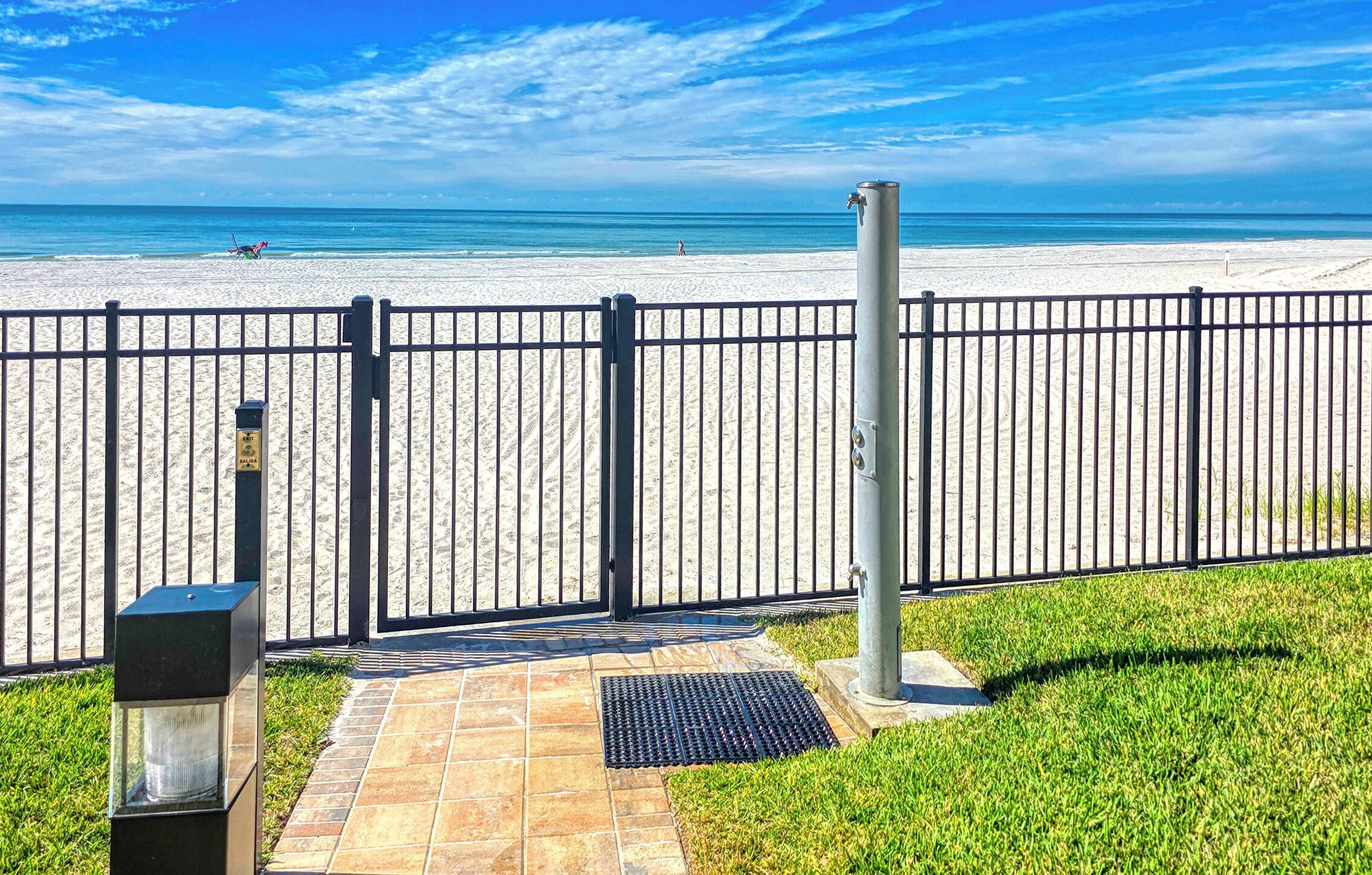 A beach view with a black metal fence, a paved walkway, and a grassy area.