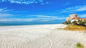 Una playa con arena, cielo azul despejado y un edificio con varios balcones con vista al mar.