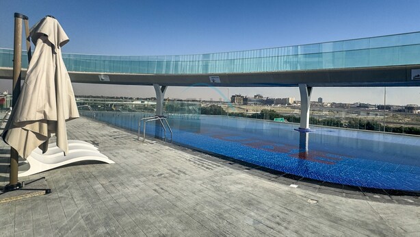 Una piscina infinita en la azotea con un puente de cristal por encima, rodeada por una terraza de madera y una zona de estar techada.