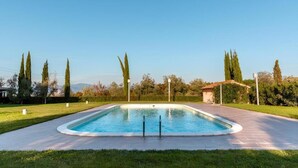 A rectangular swimming pool with a paved deck, surrounded by grass and trees, with a clear sky above. - Agriturismo gli Archi (Fauglia)