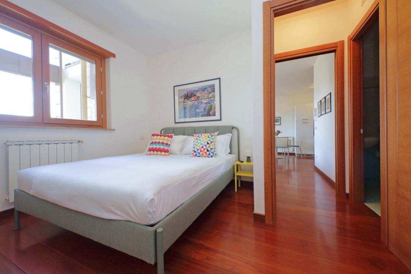 Een slaapkamer met een bed, een radiator, een raam, een schilderij en een gang.