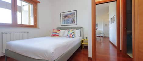 Een slaapkamer met een bed, een radiator, een raam, een schilderij en een gang.