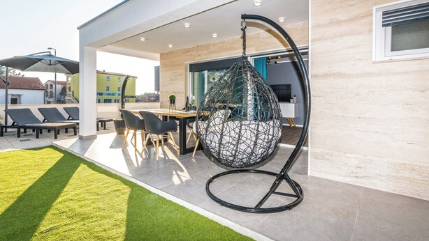 Een modern buitenterras met een hangstoel, een eethoek en een verzorgd gazon.