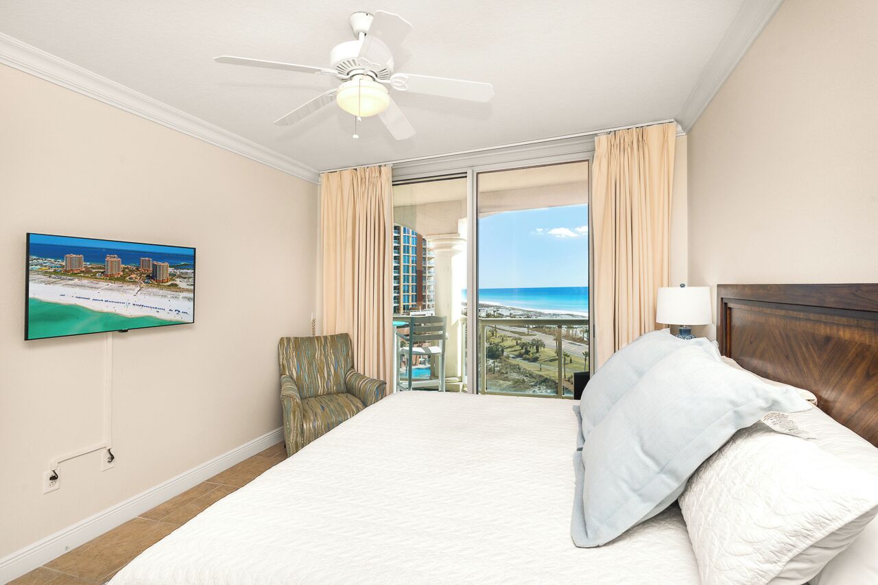A hotel room with a bed, a ceiling fan, a TV on the wall, and a balcony with a view of the beach and buildings.