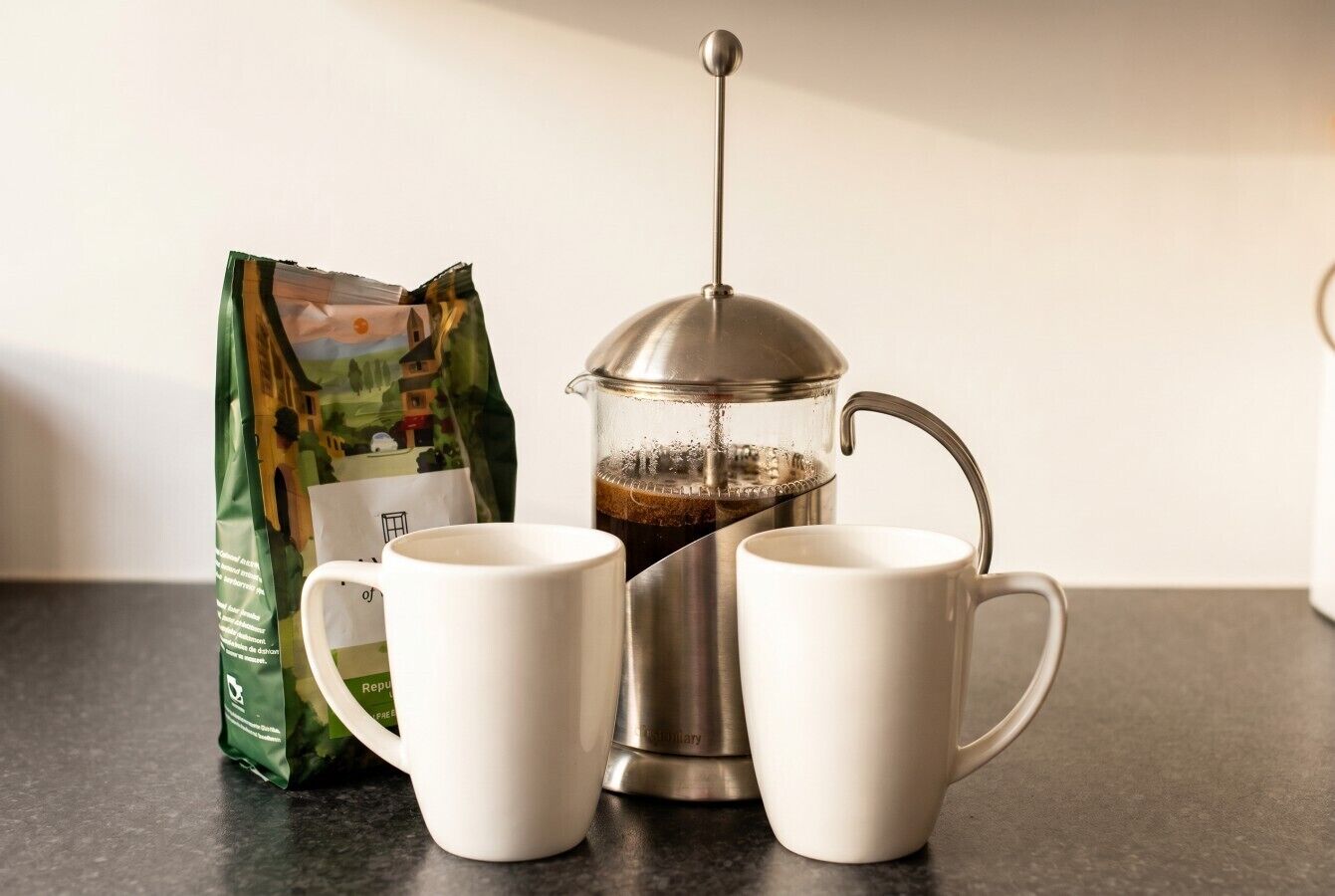 Een French press koffiezetter, twee witte mokken en een zak koffie op een aanrecht.
