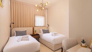 Een hotelkamer met twee bedden, een nachtkastje met een lamp, een raam met zonwering en een tafeltje met een vaas.