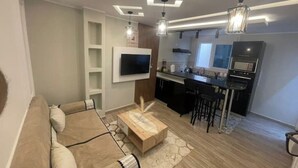 Un salon moderne avec un canapé, une table basse et un téléviseur fixé au mur. Il y a également un coin cuisine avec une table à manger et des chaises.